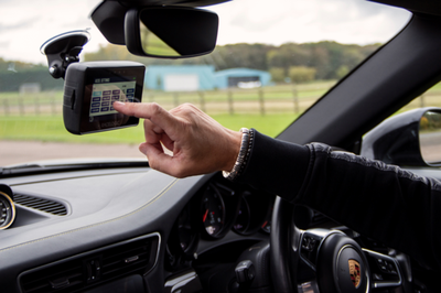 Interior view of a car on a track, showing a driver reaching toward a Performance Box Touch mounted near the rearview mirror, with dashboard and steering wheel visible.