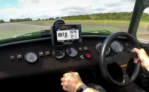 Interior driver’s view of a car on a track, showing hands on the steering wheel, dashboard controls, and a mounted Performance Box Touch displaying speed and performance data.