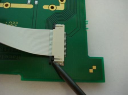Small screwdriver pushing on the tabs at the left-hand side of the ribbon cable to disconnect it from the PCB.