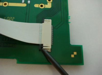 Small screwdriver pushing on the tabs at the left-hand side of the ribbon cable to disconnect it from the PCB.
