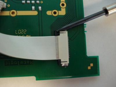 Small screwdriver pushing on the tabs at the right-hand side of the ribbon cable to disconnect it from the PCB.